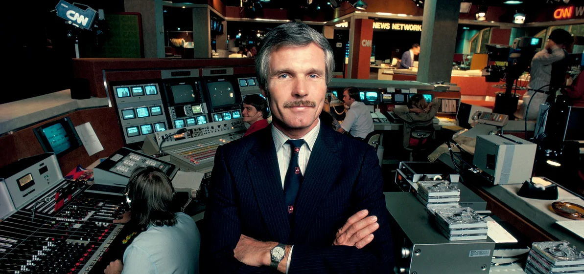 Ted Turner wearing a black suit and tie with his arms crossed standing in a media studio